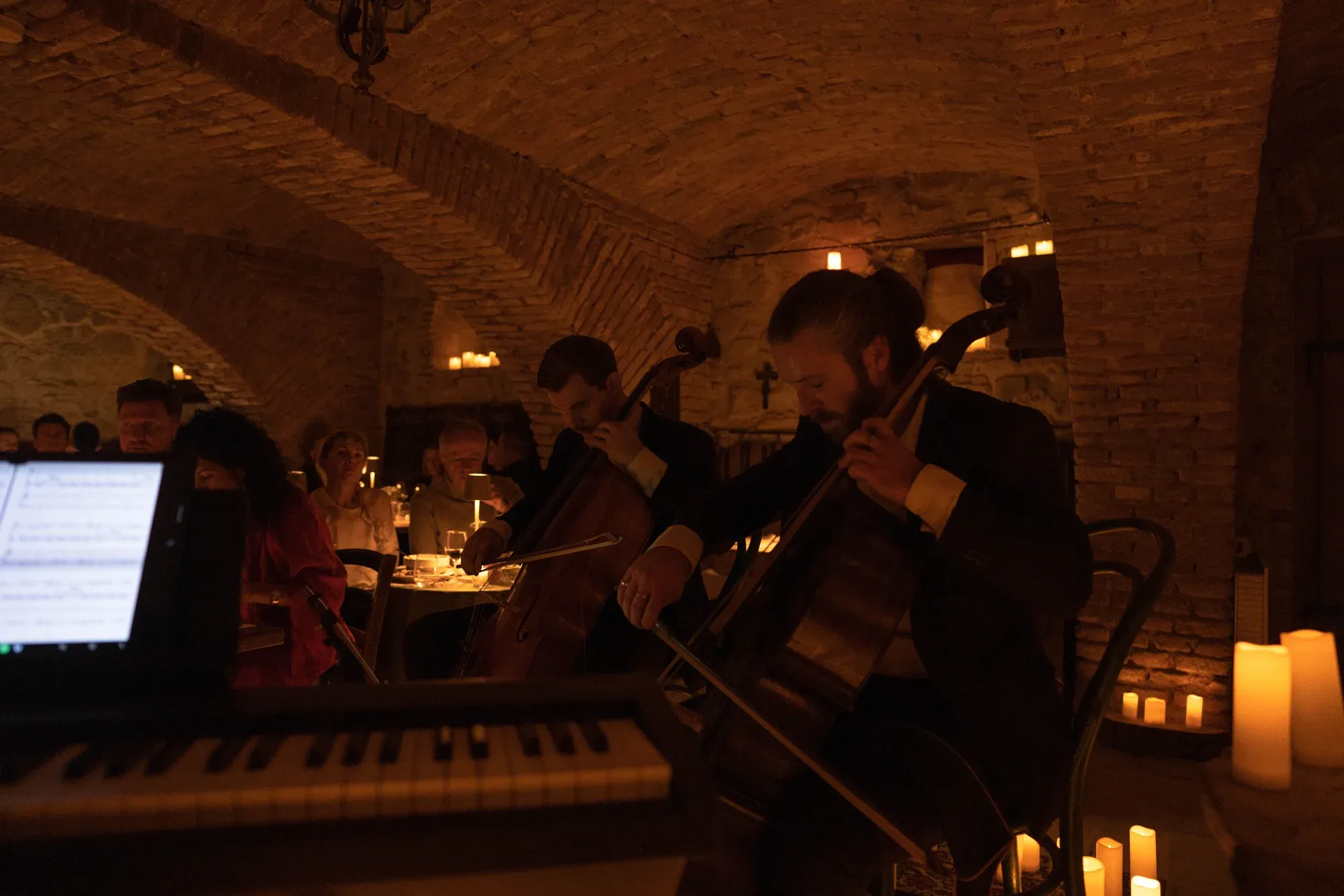Two cellists performing beside a piano in the candlelit castle wine cellar