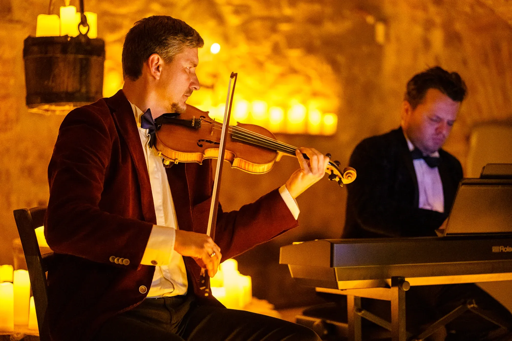 A violinist playing beside a pianist, surrounded by candlelight in the castle wine cellar